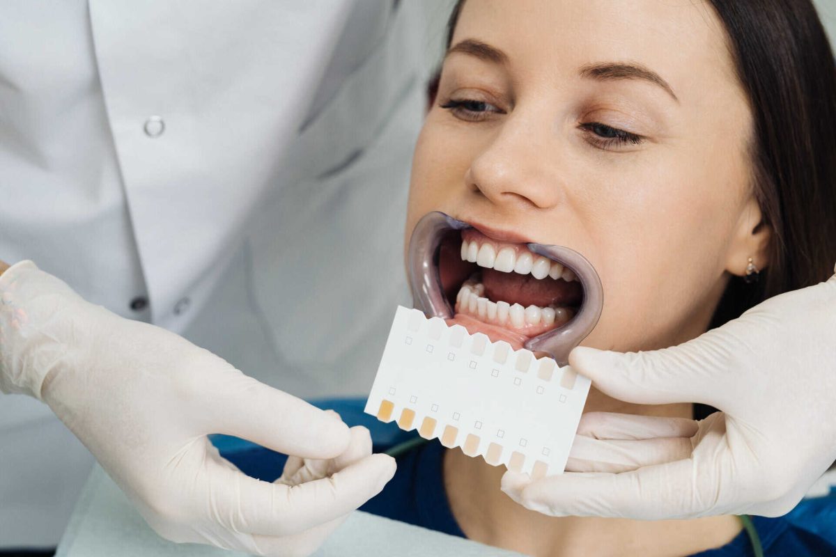 Close up portrait of beautiful young lady sitting in dental chair while stomatologist hands in sterile gloves holding tooth samples. She is smiling
