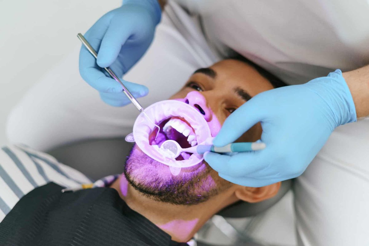 Young man with patient bib on a dental chair, pink light oriented on his teeth and a dentist who sits next to him wearing blue medical gloves while examining oral cavity